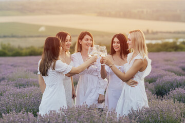Group of happy cheerful young girlfriends enjoy summer picnic in the lavender field at summer sunset and are clink with glasses with wine. Friendship concept.