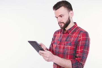 Close up photo of happy young bearded man in plaid shirt standing and using tablet isolated on white background