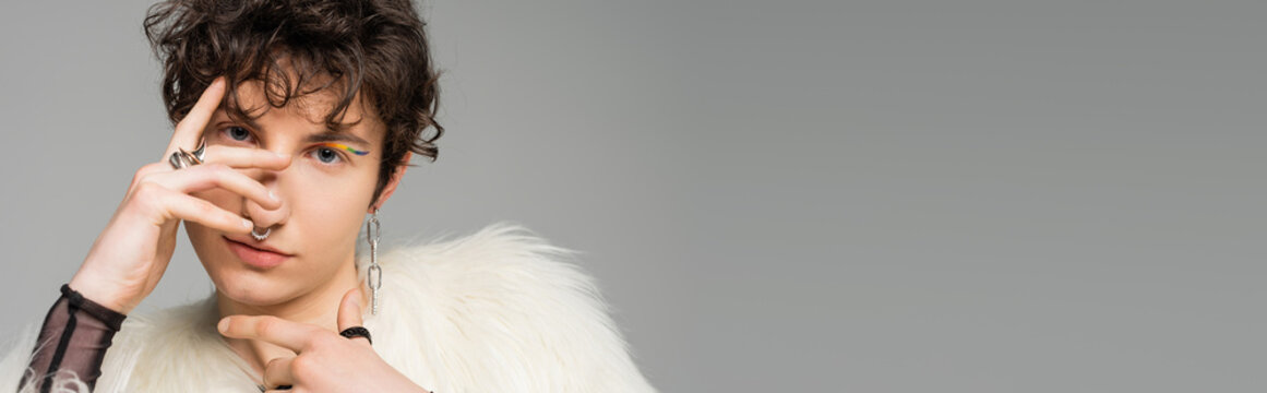 portrait of stylish bigender person in white faux fur jacket covering face with hand isolated on grey, banner.