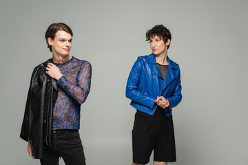 young nonbinary person in blue leather jacket looking at fashionable partner isolated on grey.