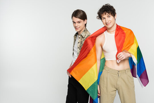 happy and stylish pangender couple with rainbow flag smiling at camera isolated on grey.