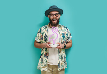A hipster guy with a beard in a hat and a Hawaiian shirt holds a neon lamp in the shape of a flamingo in his hands. On a blue studio background