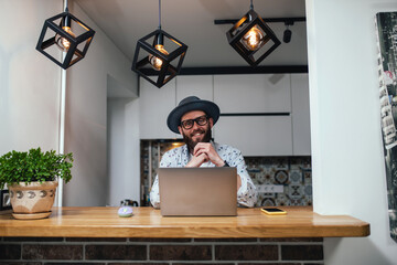 Charming male freelancer with a beard in casual clothes and a hat works at a laptop at home at a table in the kitchen, communicates with a client via the Internet.