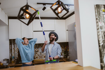 A young couple is looking at the ceiling and screaming while holding a mop because the upstairs...