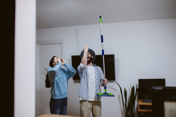 A young couple is looking at the ceiling and screaming while holding a mop because the upstairs neighbor is having a party with loud music or doing repairs in the apartment, workers are drilling with - obrazy, fototapety, plakaty