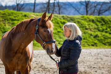 Fototapeta premium Woman stands in front of her horse in the riding arena and looks at it lovingly..