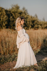 Fototapeta na wymiar A young expectant mother in the third trimester of pregnancy in a white dress hugs her stomach against the backdrop of a natural landscape. The concept of future motherhood.