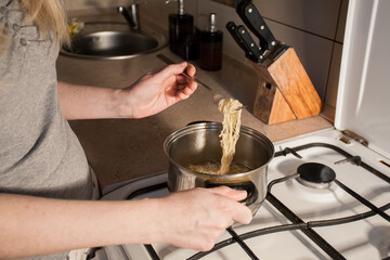 Hot instant pasta is cooked in a saucepan on the stove in the kitchen. The girl stirs the food in the bowl with a fork. Image for your creative decoration and design.