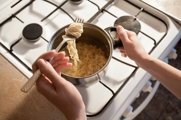 Hot instant pasta is cooked in a saucepan on the stove in the kitchen. The girl stirs the food in the bowl with a fork. Image for your creative decoration and design.