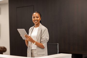 The young African-American female receptionist stands behind the reception desk with a digital tablet in her hands and smiles at the camera. - obrazy, fototapety, plakaty