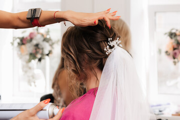 A hairdresser makes an elegant hairstyle for styling a bride with a white veil in her hair in the salon
