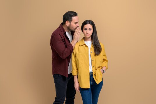 Shocked girlfriend with raised eyebrows listening to boyfriend whispering secret into her ears. Young couple discussing private gossips while standing over beige background