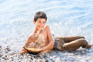 Child on beach is playing with homemade toy wooden boat with sail. Turquoise sea water all around. Sea adventures and travel concept.