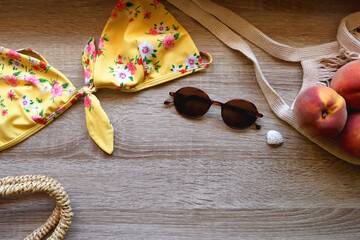 Floral yellow bikini, wicker bag, retro sunglasses, seashell, bag with peaches and cut watermelon on wooden background. Top view, copy space.
