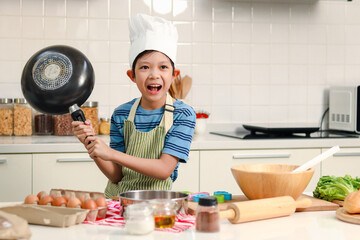 Portrait of happy Asian boy wearing apron and chef hat, holding frying pan and shouting, kid...