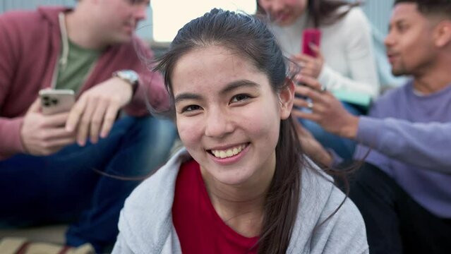 Closeup face portrait of young asian girl smiling at camera with diverse friends using smartphone device on the background. Student people enjoying time watching social media videos on cellphone.