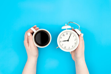 White alarm clock and a cup of coffee in female hands on a blue background, top view, morning, creative concept.