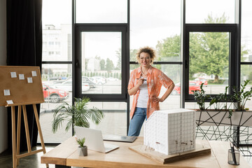 Fototapeta na wymiar happy architectural designer standing with hand on hip near gadgets and residential house model on desk.