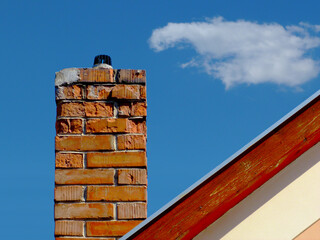 Isolated damaged clay brick chimney with weathered and spalling surface. wood trim on house gable end wall. metal flashing. white stucco exterior elevation. strong shadow. blue sky. fluffy white cloud