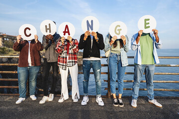 Youth Advocating for Change: Multiracial young people cover faces with "CHANGE" discs by a railing, sea, and rocks, symbolizing a call for societal transformation