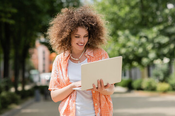 curly freelancer woman smiling while holding laptop and working remotely in park.