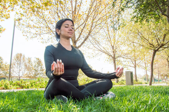 Young woman meditating in lotus or easy pose (padmasana or sukhasana) on the grass in the park, balance, work, life and peace of mind concept - image with copy space