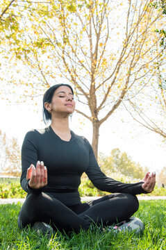 young woman meditating in lotus or easy pose (padmasana or sukhasana) on park grass; balance; work; life and peace of mind concept - image with copy space; vertical photo