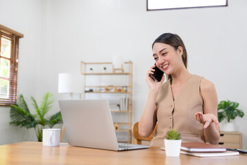 Young asian woman working with laptop computer and talking on the phone.Asian woman relaxing at work sending voice messages on mobile phone during break from work from home.