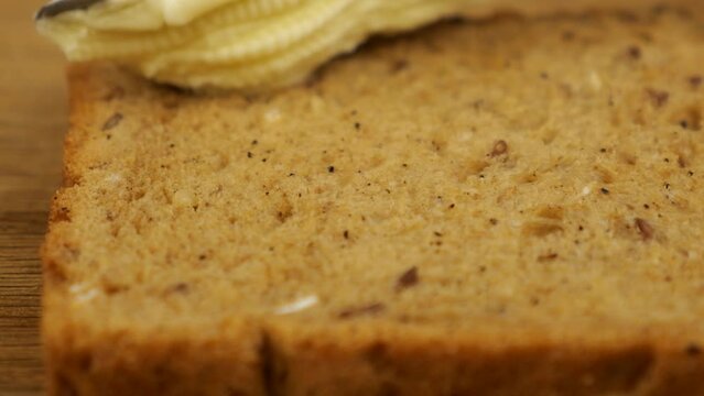 Butter is spread with a knife on a piece of multigrain bread on a wooden cutting board.
