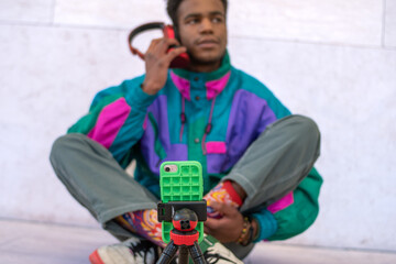 Young black man in colorful dress making self videos for social media, using tripod and mobile.