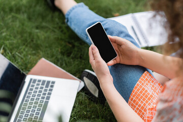 cropped view of young woman holding smartphone with blank screen while sitting near laptop and notebook on grass.