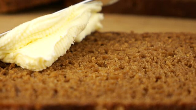 Butter is spread with a knife on a piece of rye bread on a wooden cutting board.