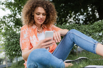 low angle view of positive freelancer woman with curly hair using smartphone in park.
