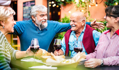 Genuine retired people having fun drinking red wine at dinner party - Senior friends eating together outside at restaurant - Dinning life style concept on vivid filter - Focus on bearded hipster man