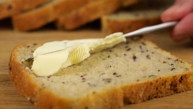 Butter is spread with a knife on a piece of multigrain bread on a wooden cutting board.