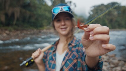 Woman angler on the river. Young woman holds the fishing fly in her hand and shows the lure to camera - 589947906