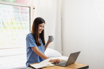 Young Asian woman with beautiful face, long hair in blue shirt sitting on white bedroom bed at home with cup of coffee and video call with laptop talking to relatives on holiday. vacation concept.