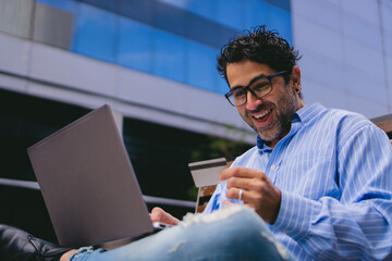 Excited middle-aged Latin man on a city bench making an online purchase with his credit card on a laptop. Copy space.