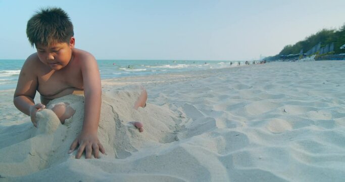 (Slow motion) A happy Asian boy who had a rather chubby figure, enjoying and playing in the sand on the sea beach in the evening sunset time.