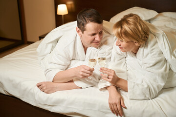 Middle-aged lady and her husband are drinking champagne in bed