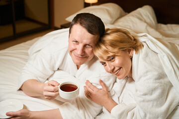 Happy couple in white bathrobes relaxing in bed