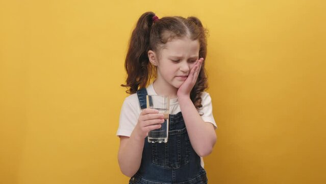 Portrait of unhealthy sad little girl kid touching chin suffering tooth ache, has sensitive teeth, feels pain after drinking cold water, posing isolated on yellow color background wall in studio