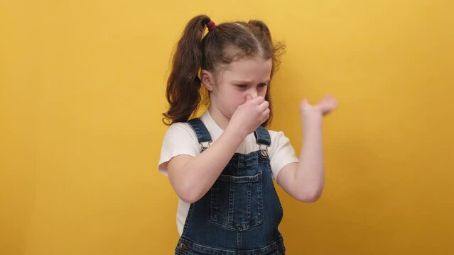 Portrait of little girl kid wearing casual clothes smelling something stinky and disgusting, intolerable smell, holding breath with fingers on nose, isolated on yellow background. Bad smell concept