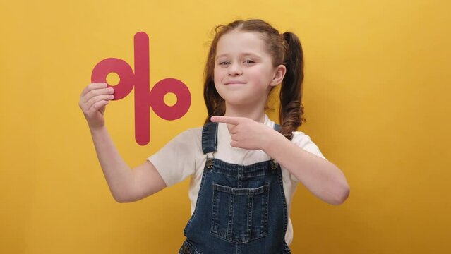 Portrait of pleasant happy little girl kid holding red percent mark, expressing positive emotions, presenting discounts, posing isolated over yellow color background wall in studio. Sale concept
