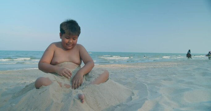 (Slow motion) A happy Asian boy who had a rather chubby figure, enjoying and playing in the sand on the sea beach in the evening sunset time.