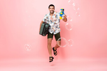 Happy handsome Caucasian tourist man in Thai summer Songkran outfit jumping with water gun and suitcase in pink color studio background