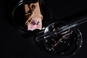 Details on a bar of soap with natural organic ecologically-friendly ingredients, wrapped in eco paper and lavender flowers with laboratory glass stick on petri dish, isolated over black background