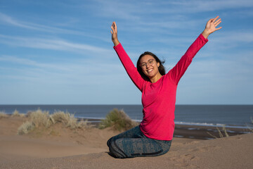 A brunette Latina woman raising her arms in happiness and sipping nonchalantly on the dunes with...