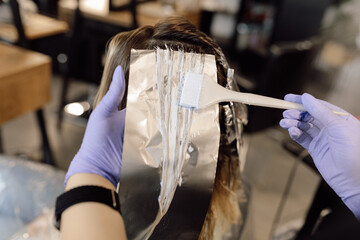 Unrecognizable woman hairstylist colorist hold strand of long hair on foil, applying hair dye with...