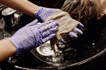 Cropped photo of colorist wearing blue gloves washing long dyed hair after coloring in black basin...